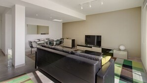 A modern living room with a black sofa, a dining area with a glass table, and a TV on a stand. - Marvellous Seafront Apartment in the Best Location (Sliema)