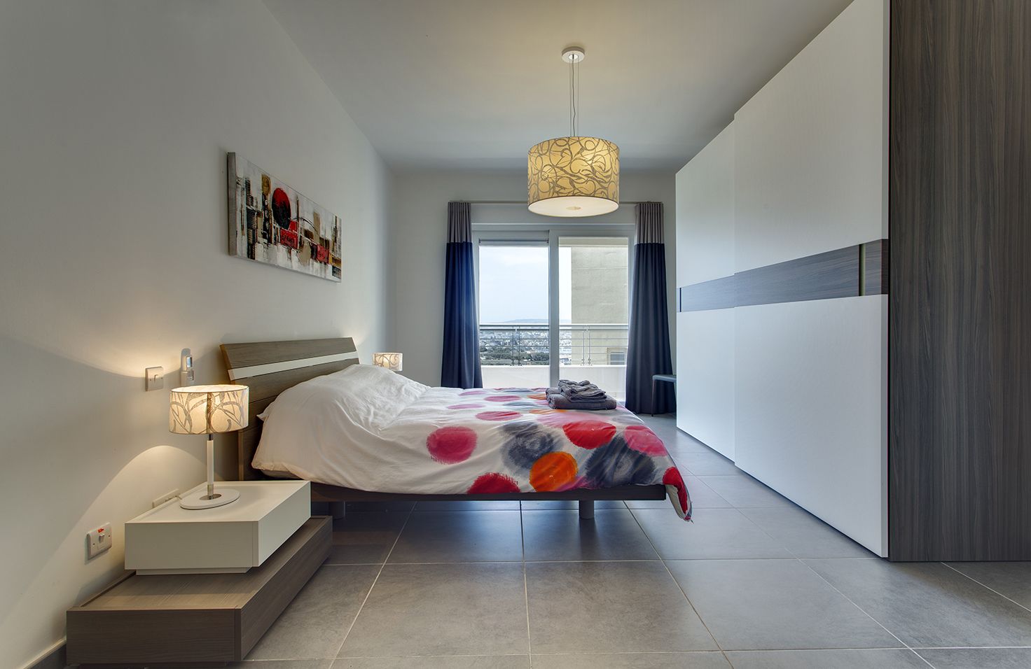 A modern bedroom with a bed, bedside lamps, a view of the city through large windows, and a balcony.