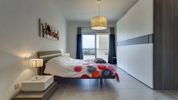 A modern bedroom with a bed, bedside lamps, a view of the city through large windows, and a balcony.