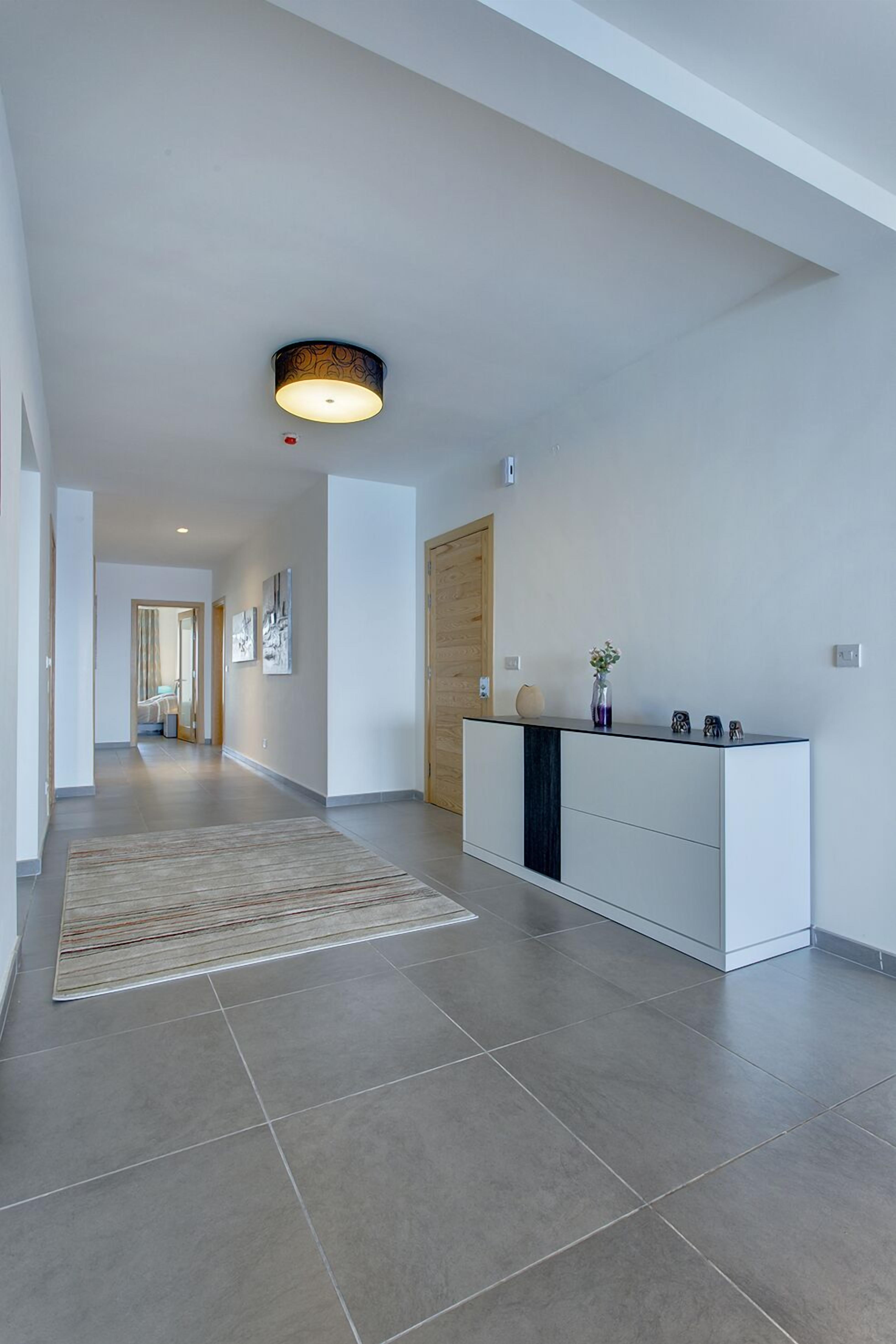Een moderne gang met een lichtgekleurde betegelde vloer, een houten deur, een witte console tafel met decoratieve voorwerpen, en een tapijt met dessin.