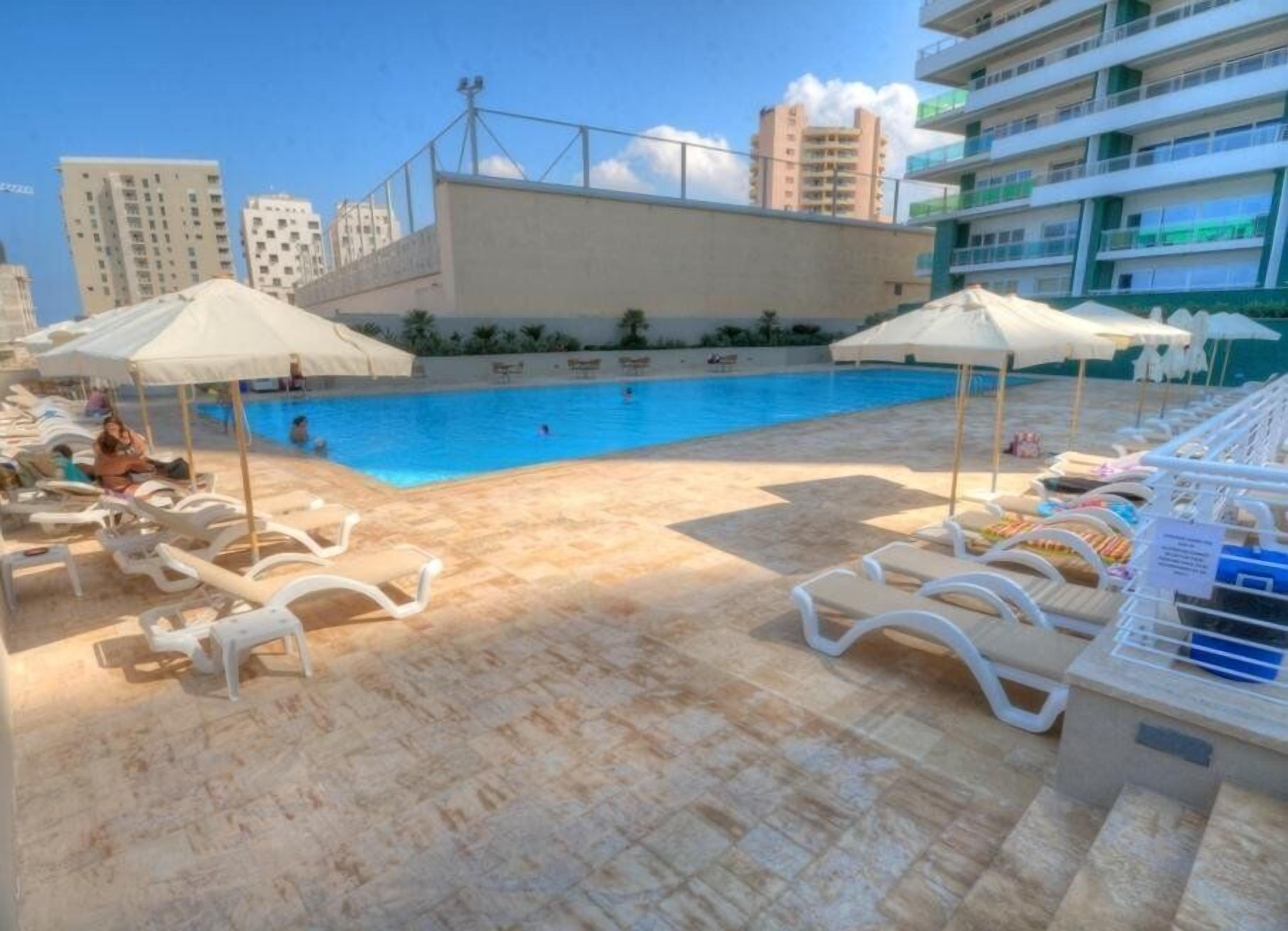 A poolside area with lounge chairs and umbrellas, a swimming pool, and high-rise buildings in the background.