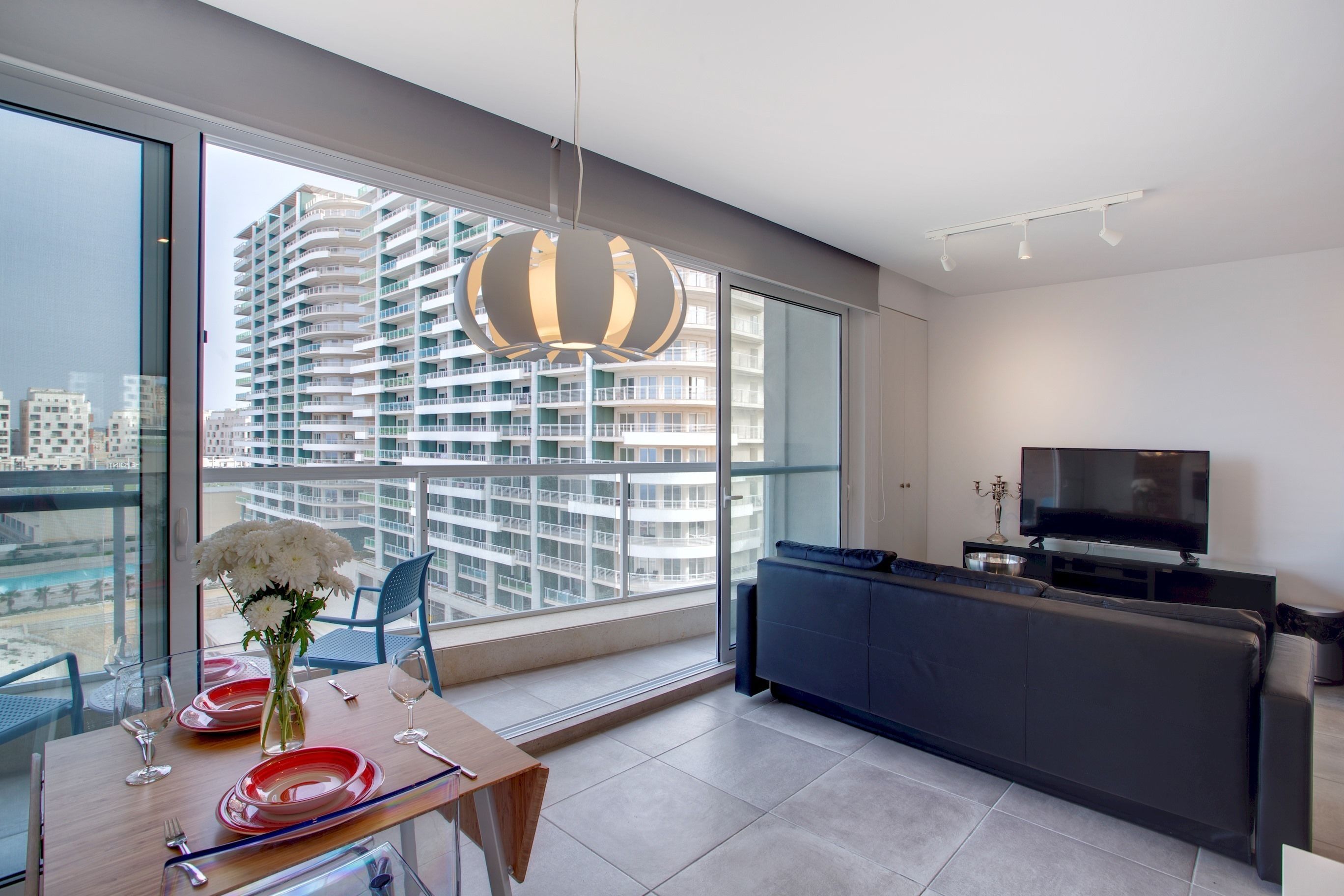 A modern living room with a large window, a sofa, a TV, and a dining area with a table set for two.