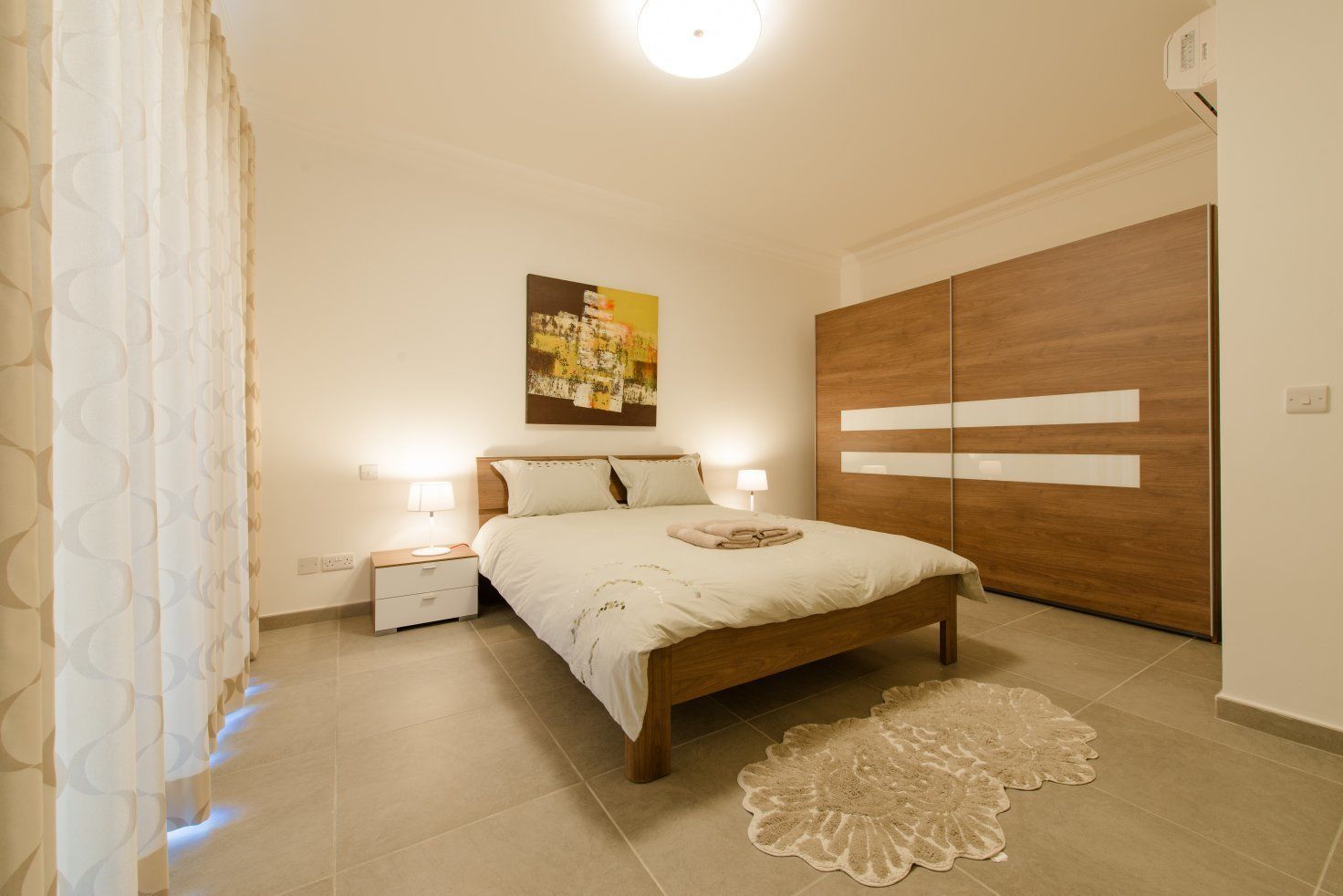 A bedroom with a bed, nightstands, a wooden wardrobe, a decorative rug, and a wall art piece.