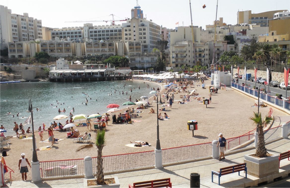 A beach with people sunbathing and swimming, a promenade with benches, and buildings in the background.