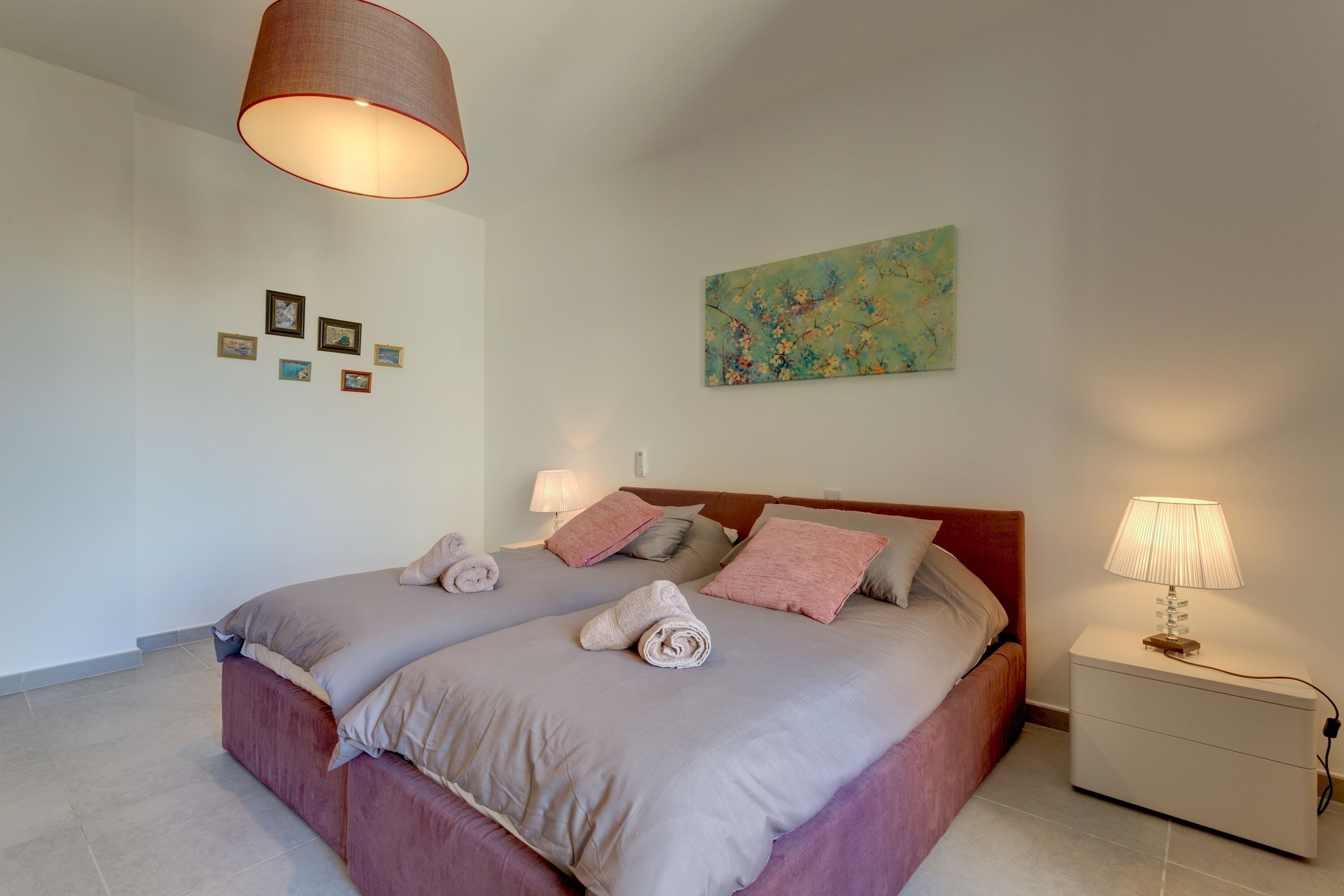 A neatly made double bed with pink and grey bedding, a bedside table with a lamp, and a wall with a floral painting and small framed pictures.