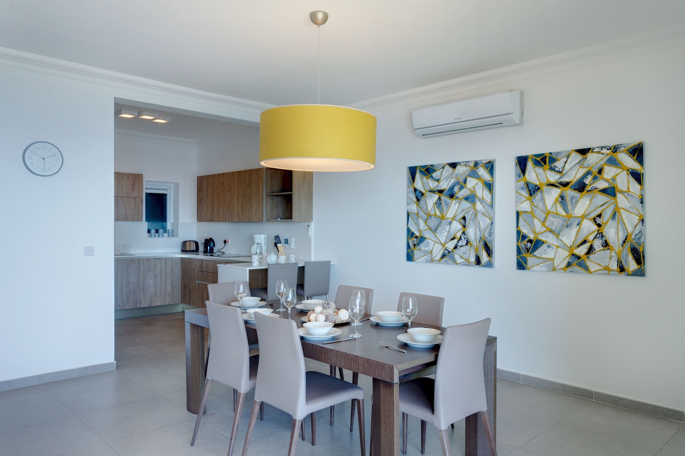 A modern dining area with a wooden table set for four, a yellow pendant light, and abstract art on the walls.