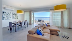 Ein modernes Wohnzimmer mit einer Couch, einem Esstisch, Stühlen, einem Balkon mit Meerblick und Pendelleuchten.
