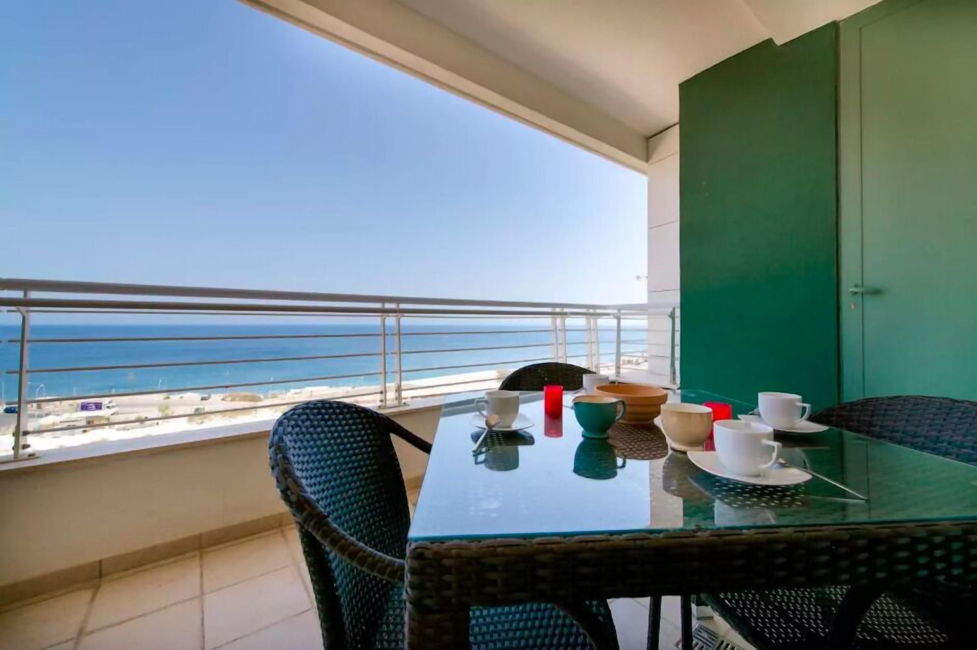 A balcony with a glass table, wicker chairs, and a view of the sea.