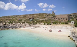 Ein Strand mit klarem türkisblauem Wasser, ein paar Sonnenbadende und ein Gebäude mit einem Turm im Hintergrund.