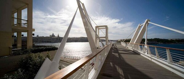A pedestrian bridge with a wooden walkway and white railings, overlooking a body of water and a distant cityscape.