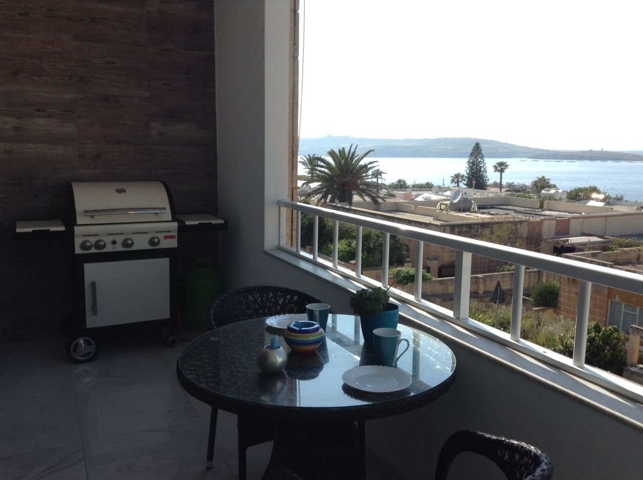 A balcony with a view of the sea, a grill, and a table with dishes and a plant.