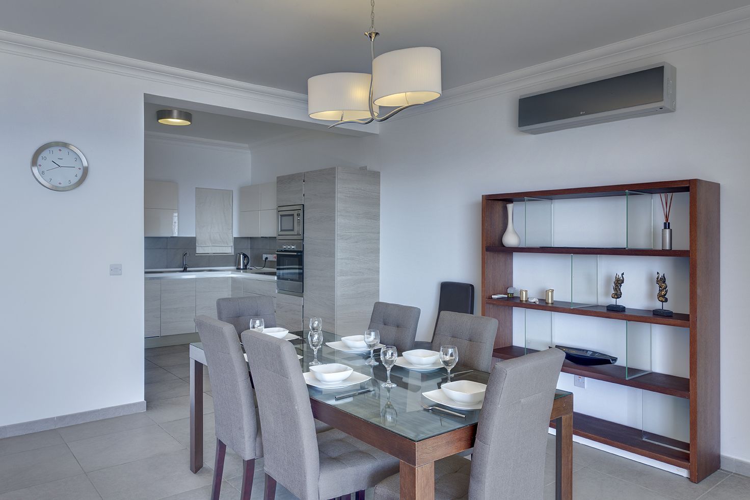 A modern dining area with a glass table, grey chairs, and a wooden bookshelf with decorative items.