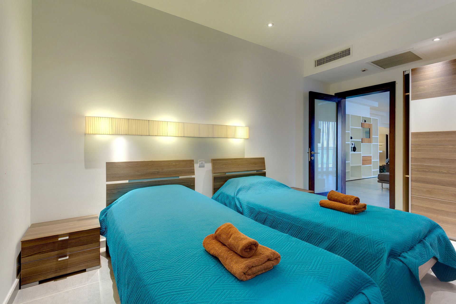 A hotel room with two beds, each with a teal bedspread and a brown towel. There is a wooden headboard, a wall-mounted light, and a door leading to a bathroom.