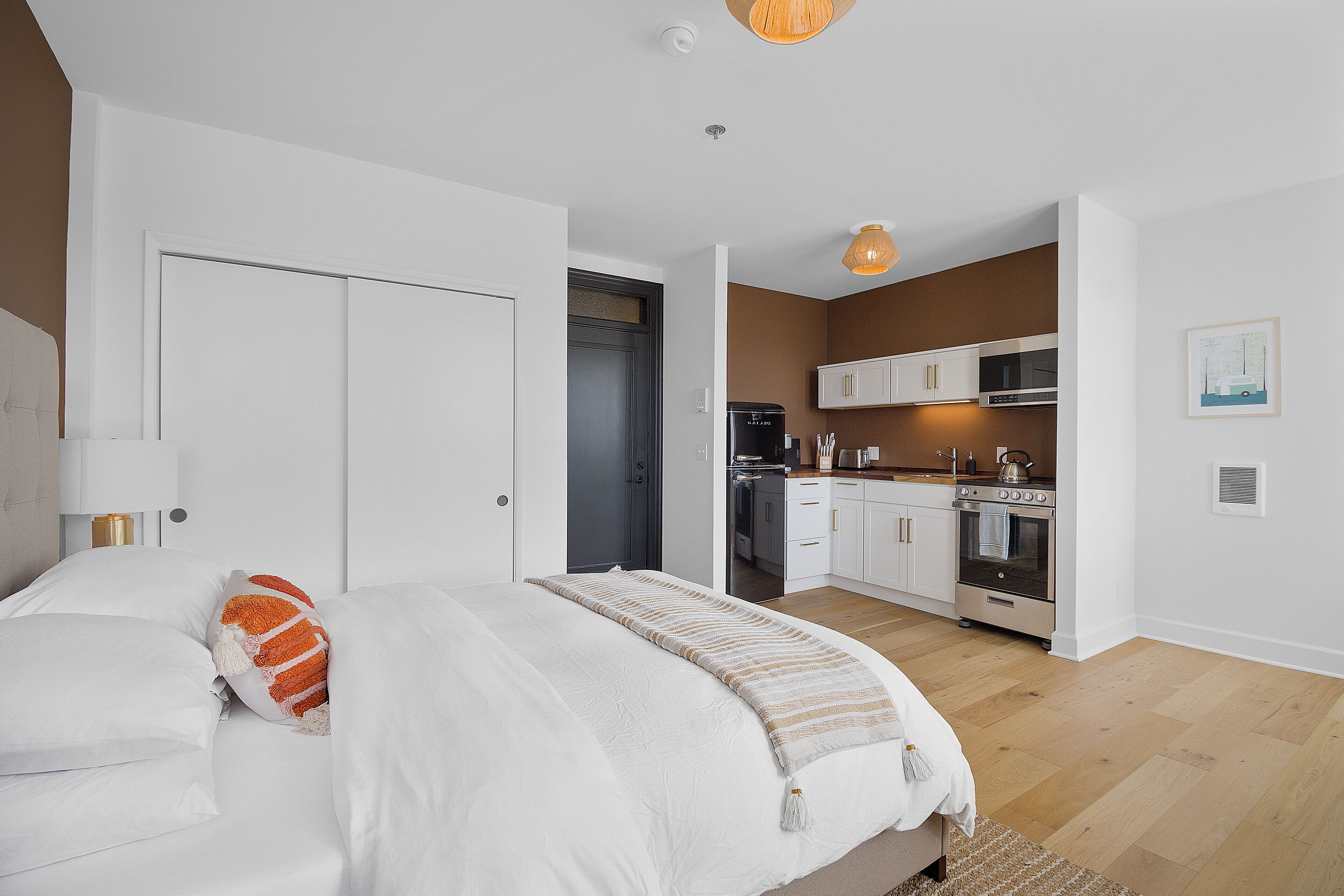 Une chambre moderne avec un grand lit, une table de chevet et une vue sur un coin cuisine équipé de placards et d’appareils électroménagers.