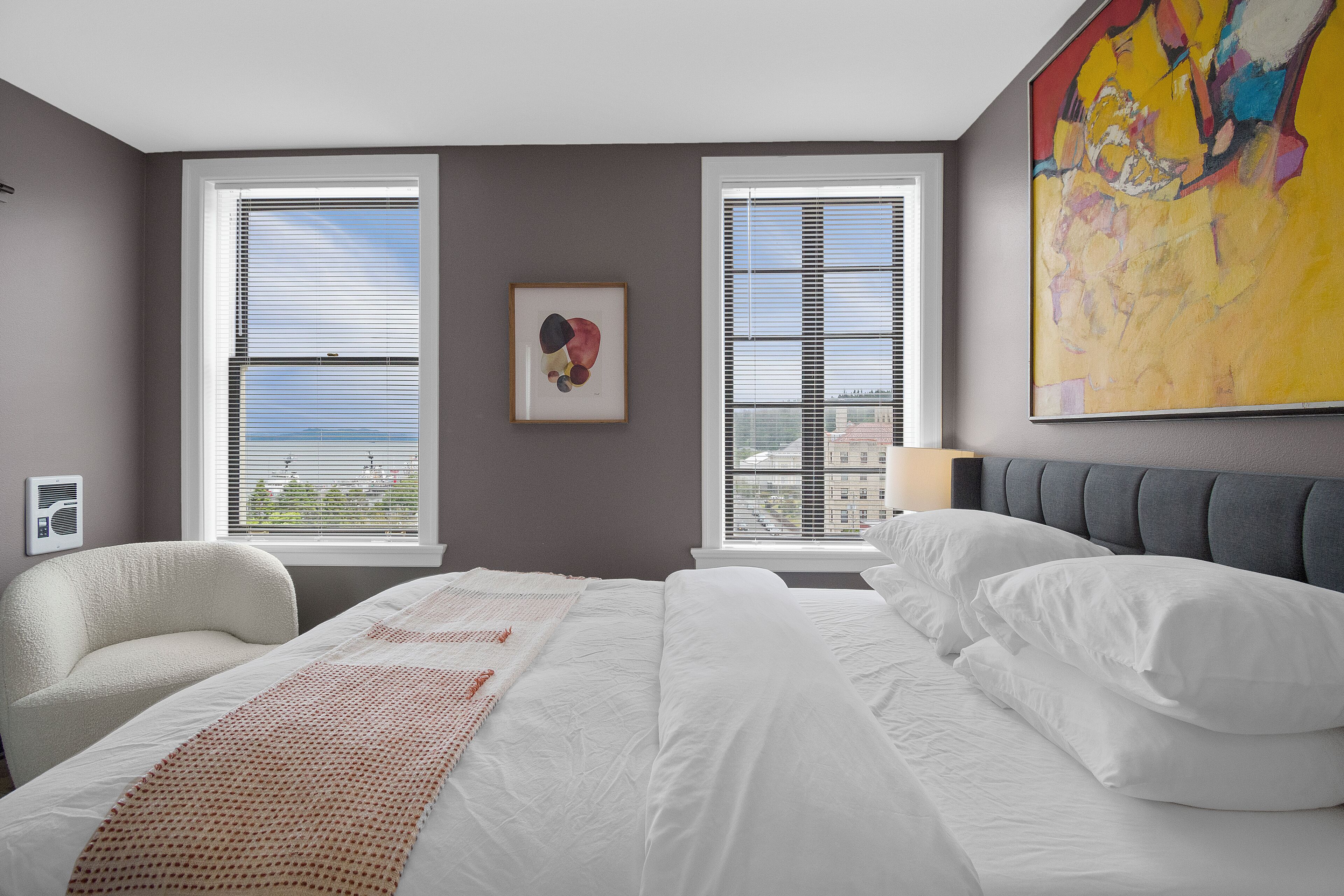A hotel room with a large bed, two windows, a chair, and a colorful painting.