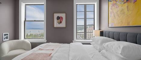 A hotel room with a large bed, two windows, a chair, and a colorful painting.