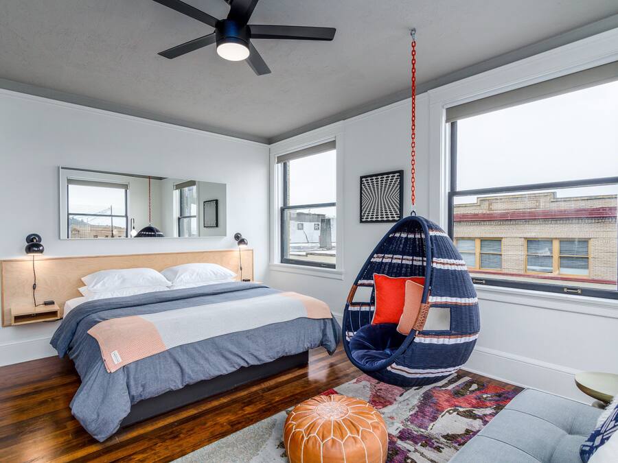 A modern bedroom with a bed, a hanging chair, a large window, and a ceiling fan.