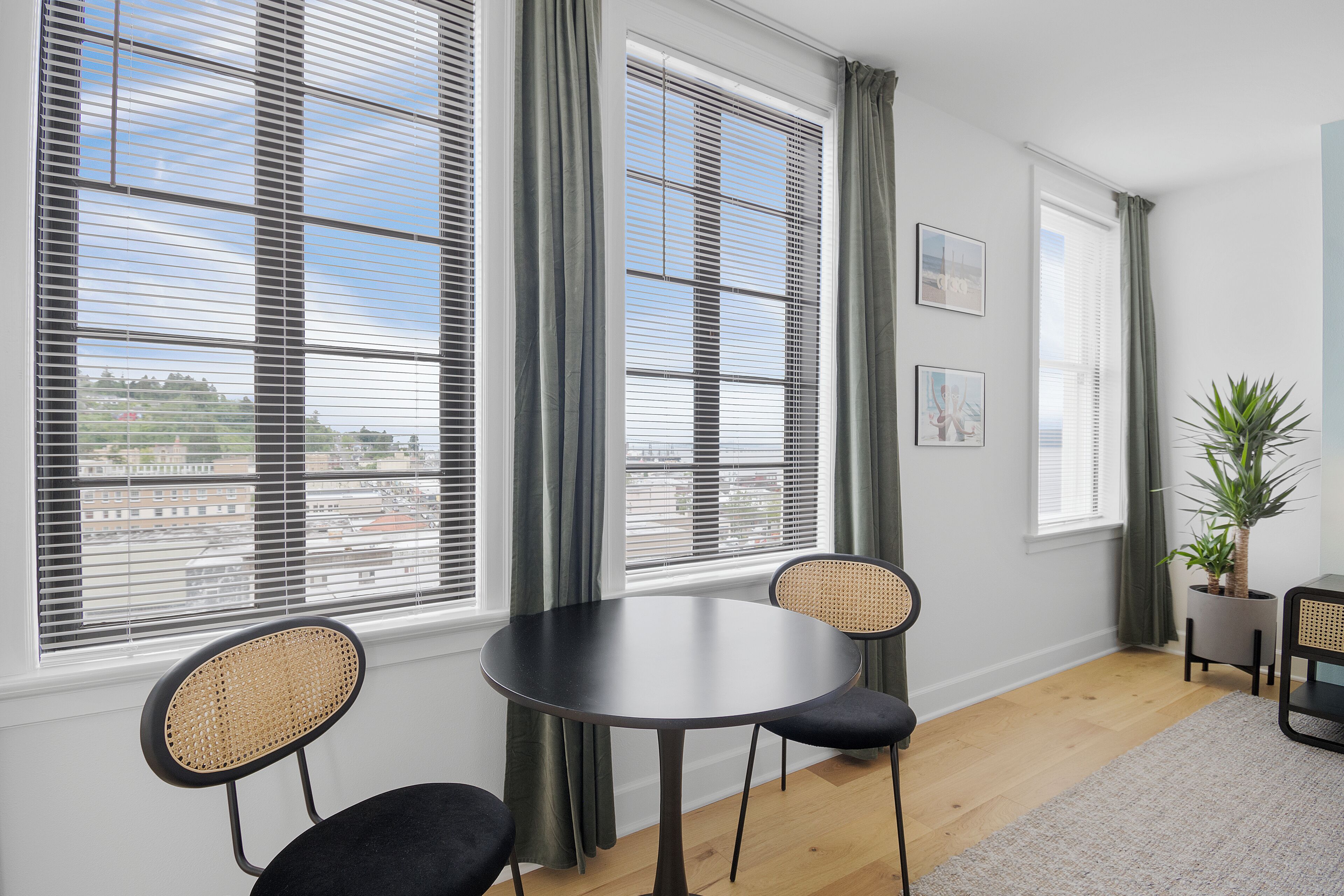 A room with a round table, two chairs, a potted plant, and large windows with blinds.