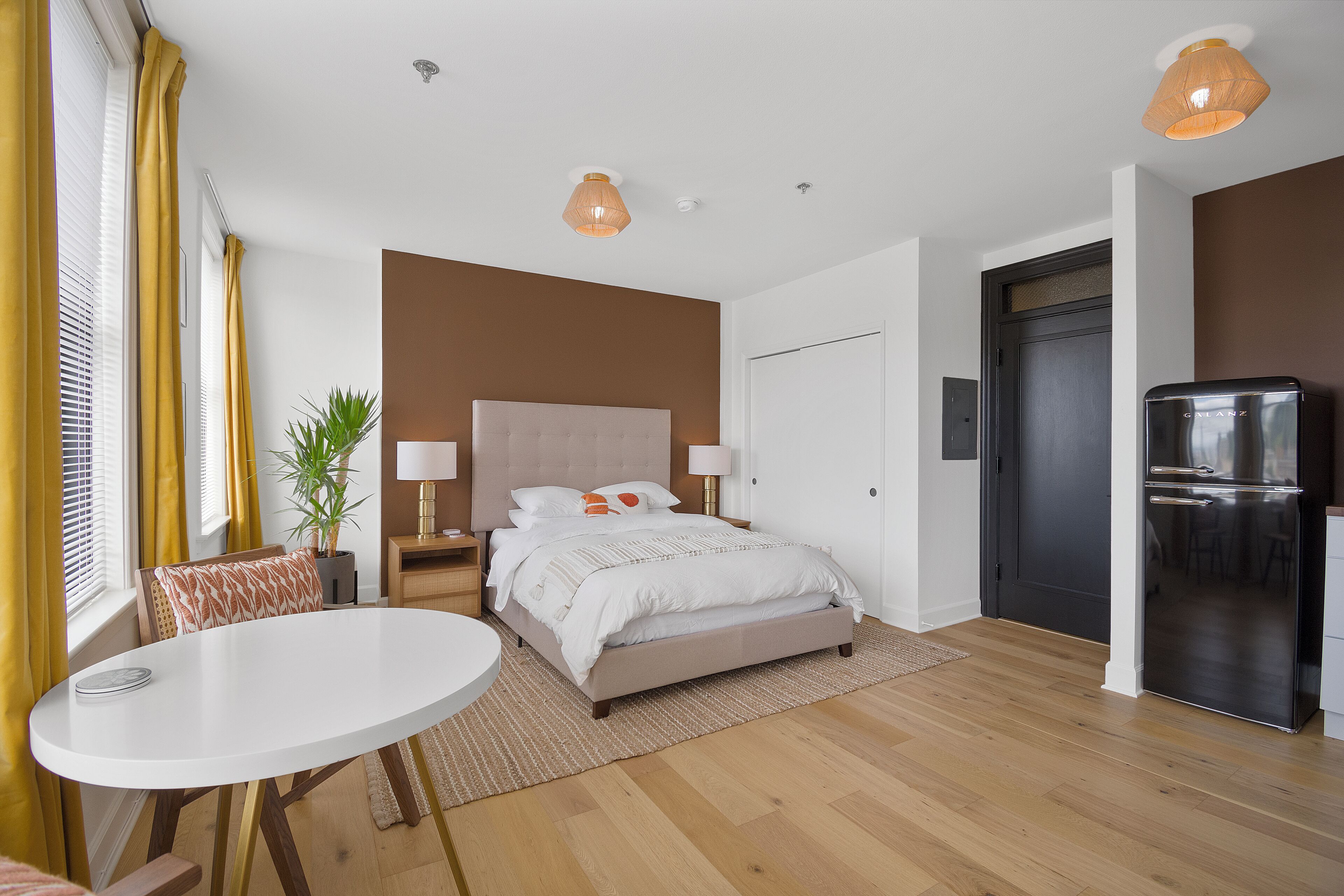 Ein modernes Schlafzimmer mit Bett, Nachttischen, einem kleinen Tisch, einer Topfpflanze und einem Kühlschrank.