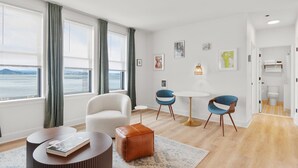 A modern hotel room with a large window offering a view of the sea, a round dining table, two blue chairs, a white armchair, and a small round table with a book. - Norblad Hotel (Astoria)