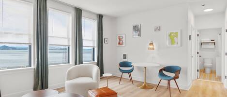 A modern hotel room with a large window offering a view of the sea, a round dining table, two blue chairs, a white armchair, and a small round table with a book.