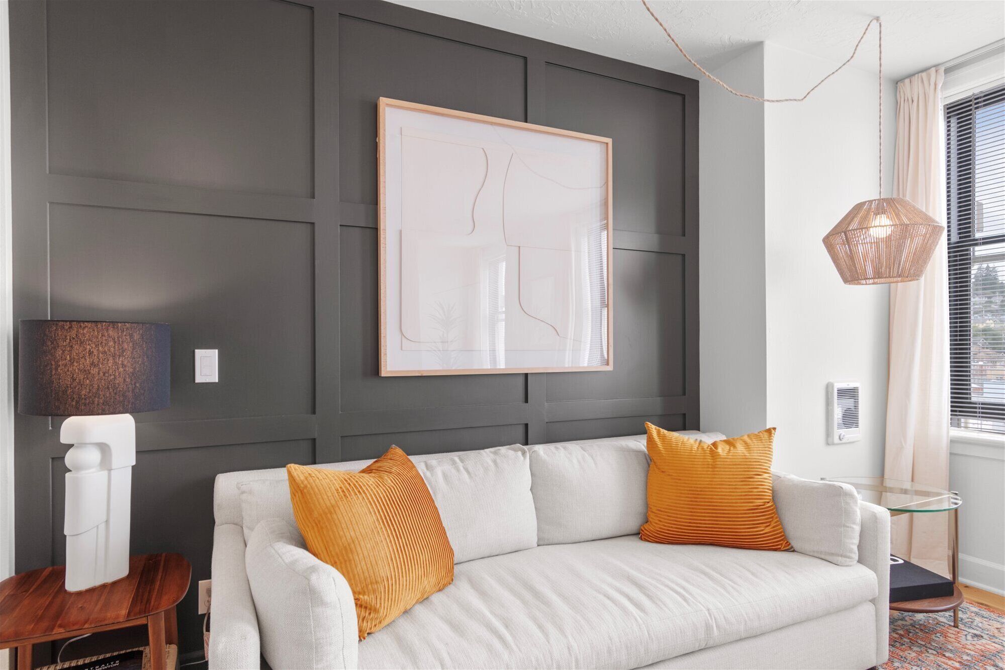 A modern living room with a grey accent wall, a framed abstract artwork, a white sofa with orange cushions, a wooden side table, and a glass coffee table.
