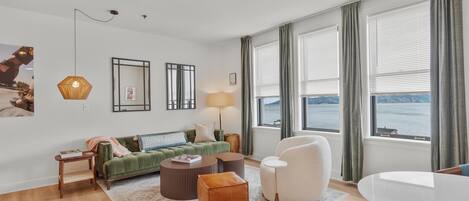 Una sala de estar moderna con un sofá verde, un otoman marrón, un sillón blanco y una mesa de centro redonda. Hay dos ventanas grandes con cortinas, una lámpara y un espejo fijado a la pared.