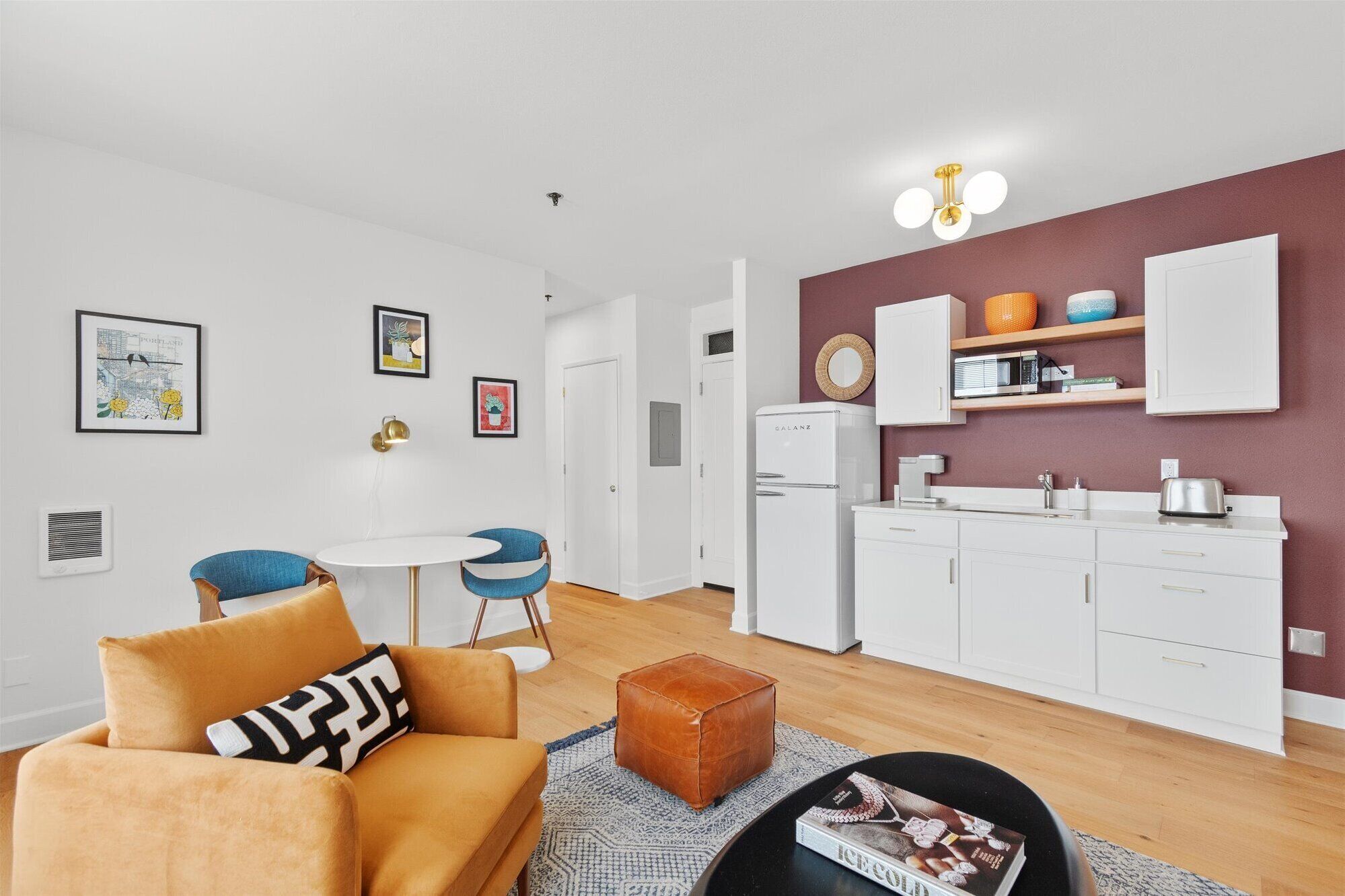 En kompakt opholdsstue med tekøkken, spiseplads med rundt bord og blå stole, samt en stue med sofa og skammel.