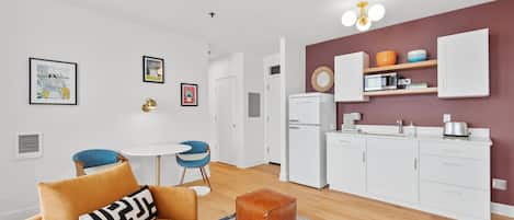 En kompakt opholdsstue med tekøkken, spiseplads med rundt bord og blå stole, samt en stue med sofa og skammel.