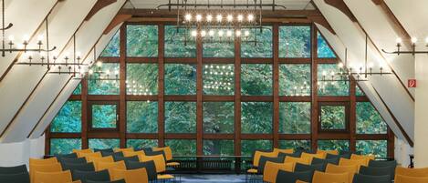 A conference room with rows of chairs, large windows, and a high ceiling with exposed beams and hanging lights.