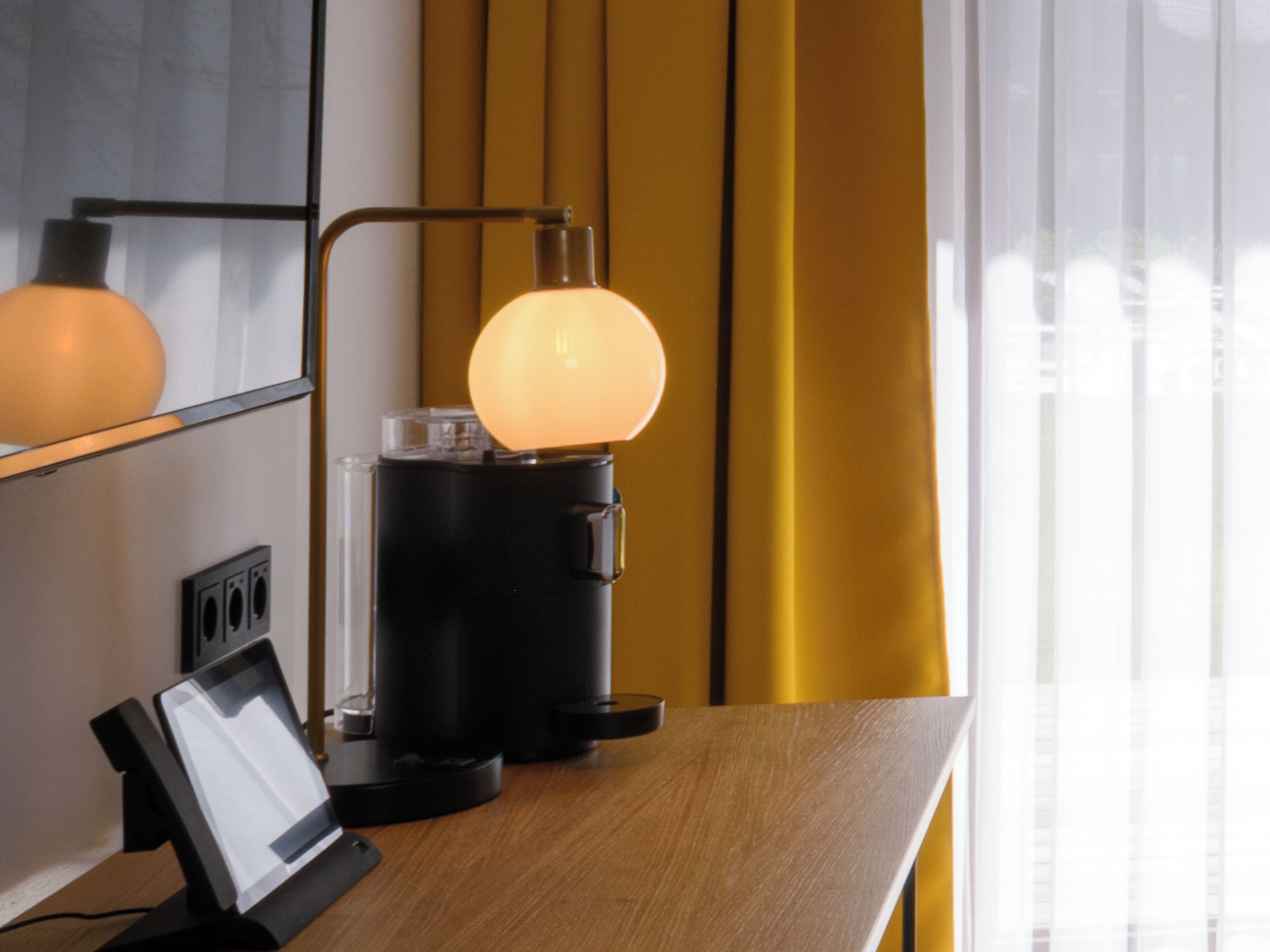 A modern hotel room with a wooden desk, a coffee machine, a lamp, and a mirror.