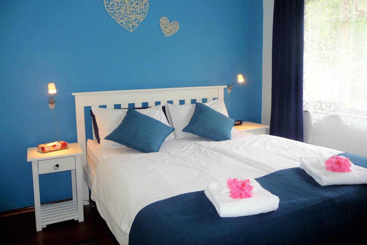 A neatly made bed with blue and white bedding, a bedside table with a lamp, and decorative heart-shaped wall art.