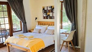 A bedroom with a bed, a chair, a mirror, and a window with curtains. - Inn2Wilderness Guesthouse (Wilderness)