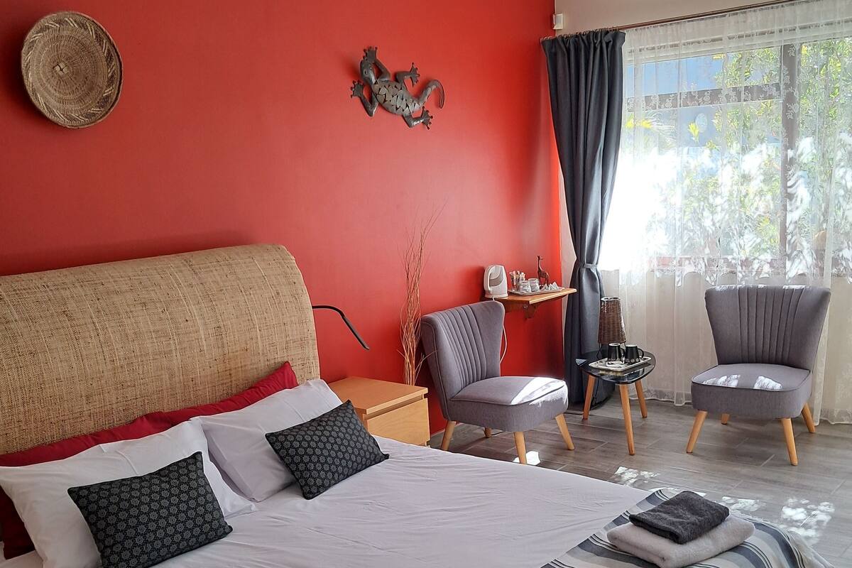 A bedroom with a large bed, a red accent wall, a small table with a lamp, and a chair.