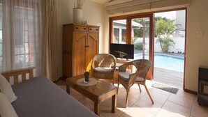 A room with a bed, a wooden table with a drink, wicker chairs, a TV, and a sliding glass door leading to a pool. - Inn2Wilderness Guesthouse (Wilderness)