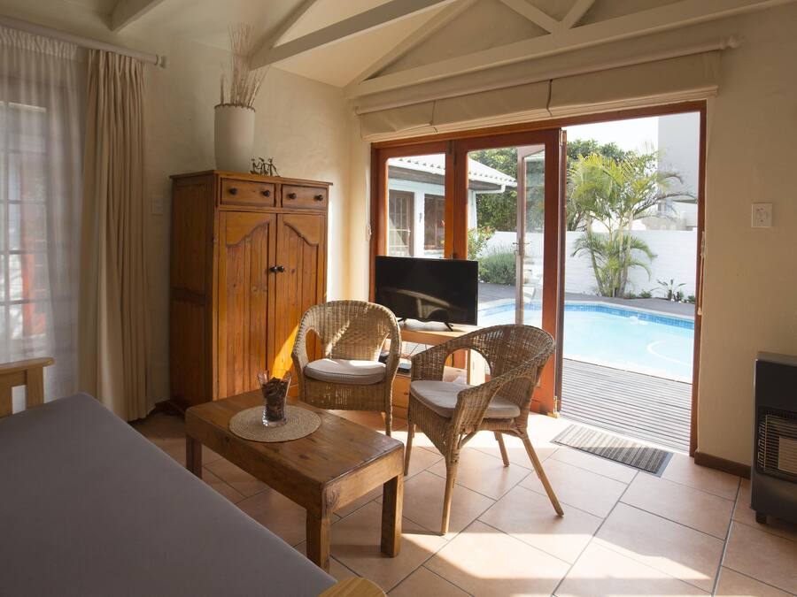 A room with a bed, a wooden table with a drink, wicker chairs, a TV, and a sliding glass door leading to a pool.