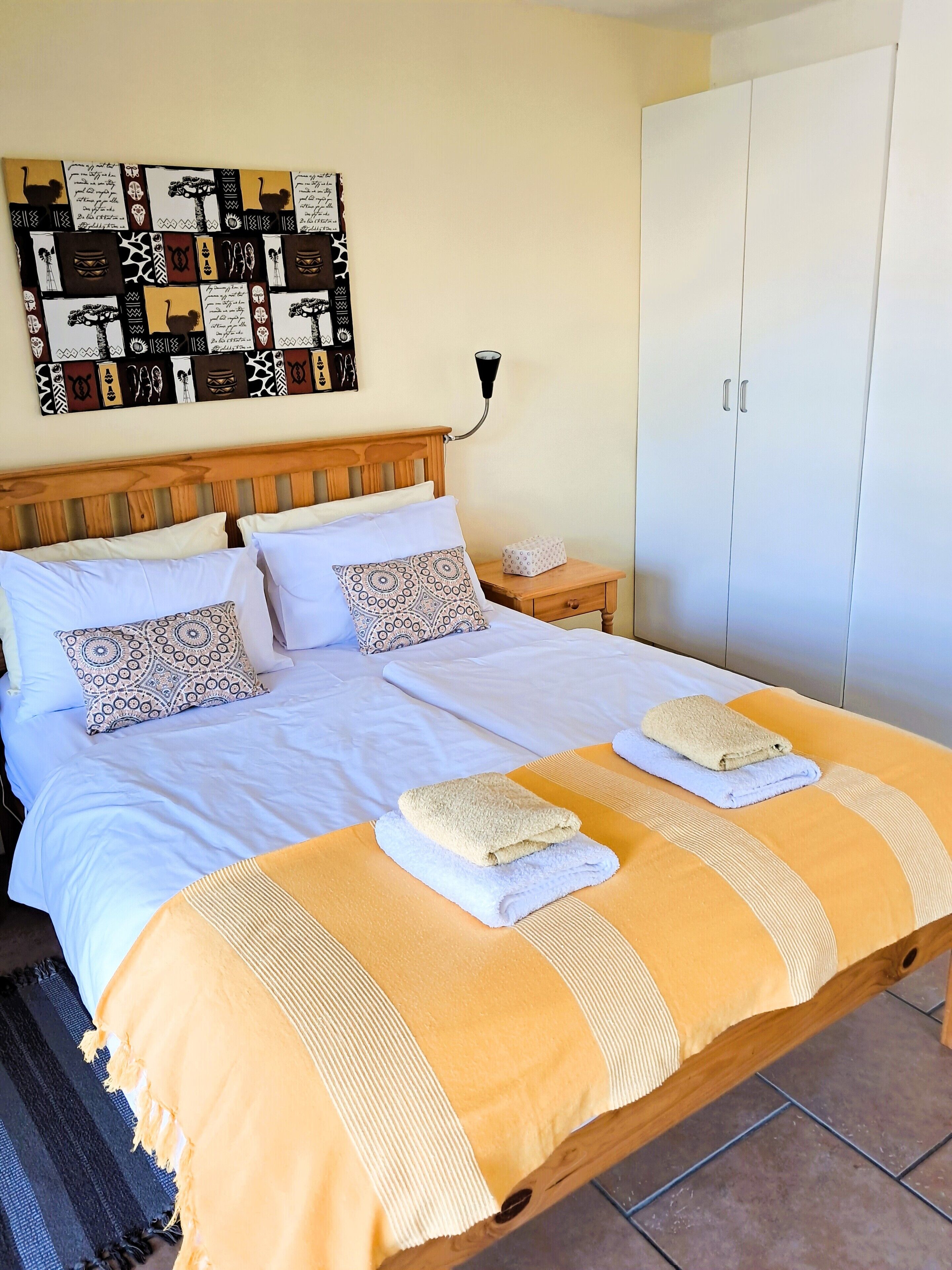 A neatly made bed with two towels, a wall with patterned artwork, a bedside table with a lamp, and a wardrobe.