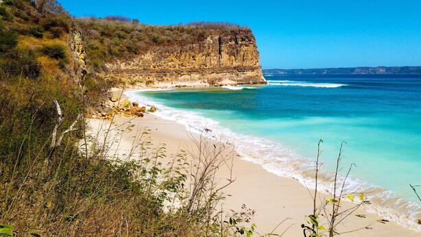 Eine Küstenlandschaft mit Sandstrand, klarem türkisblauem Wasser und einer felsigen Klippe.