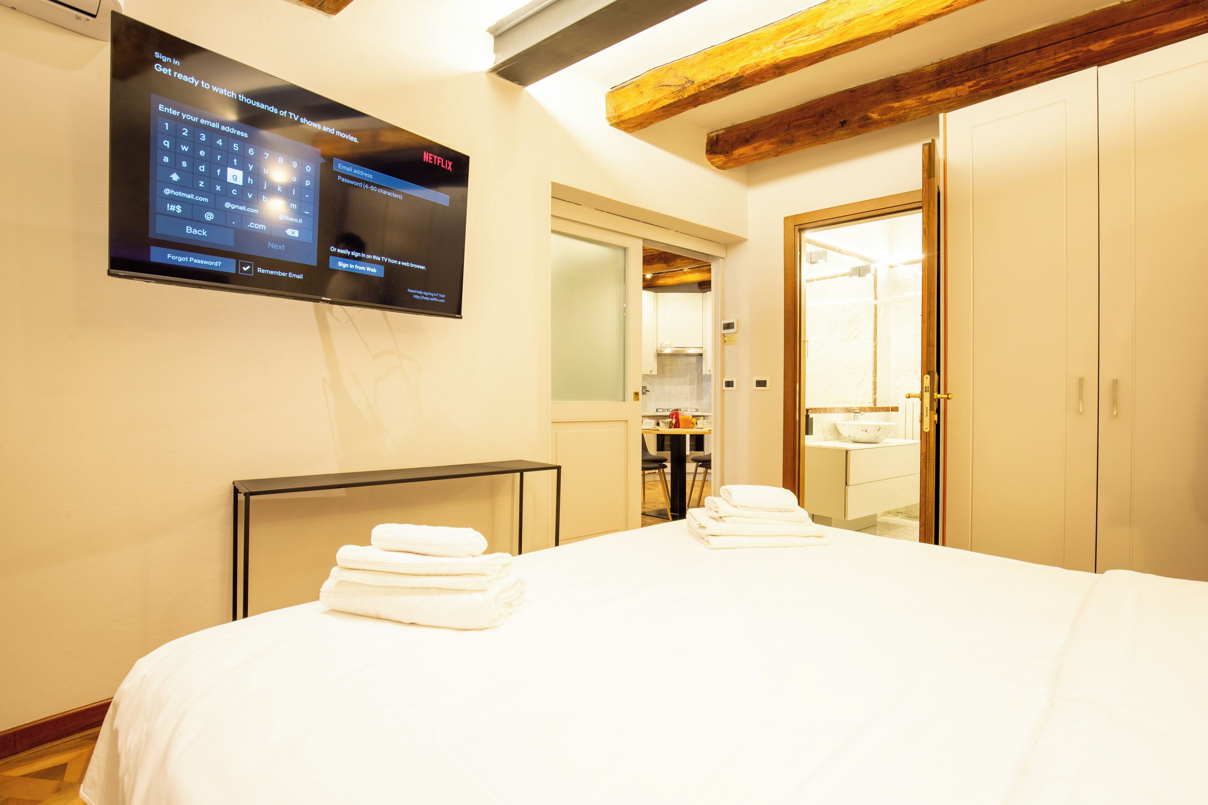A hotel room with a bed, a television mounted on the wall, a wooden ceiling, and a bathroom visible through an open door.
