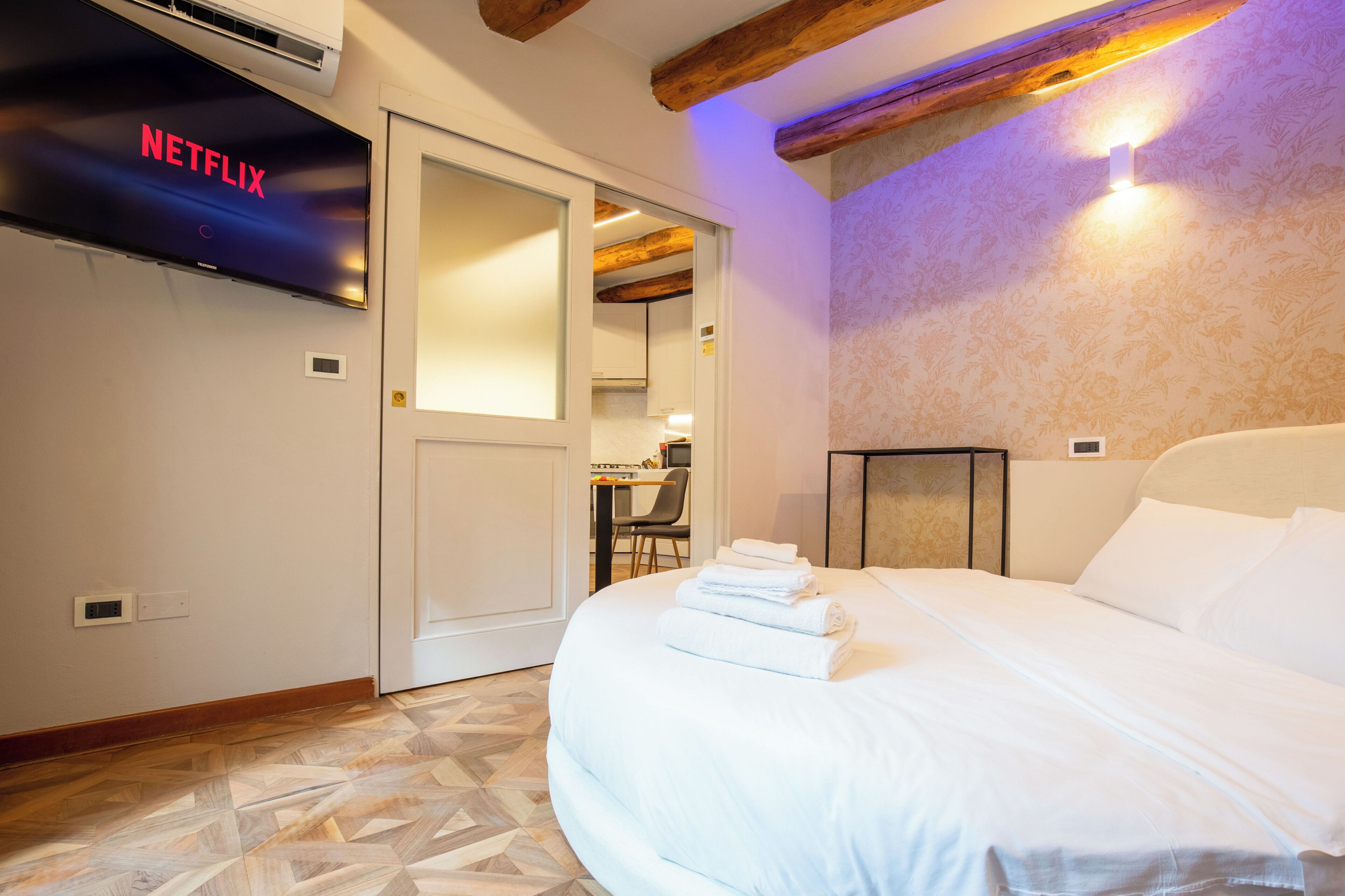 A hotel room with a bed, a flat-screen TV mounted on the wall, a wooden ceiling, and a patterned wall.