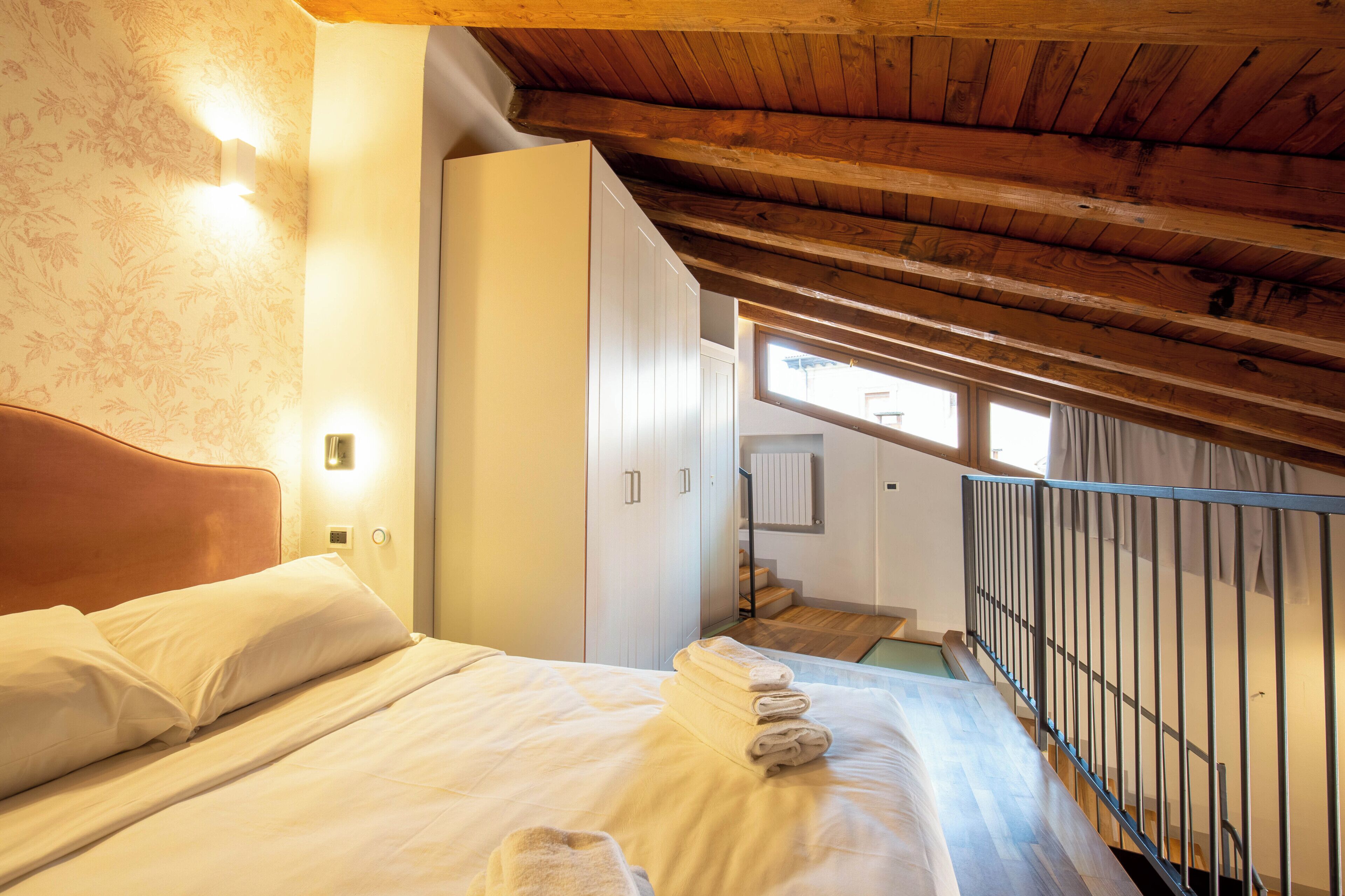 A bedroom with a wooden ceiling, a bed with white bedding, a nightstand, a wall-mounted light, and a view of a staircase leading to an upper floor.