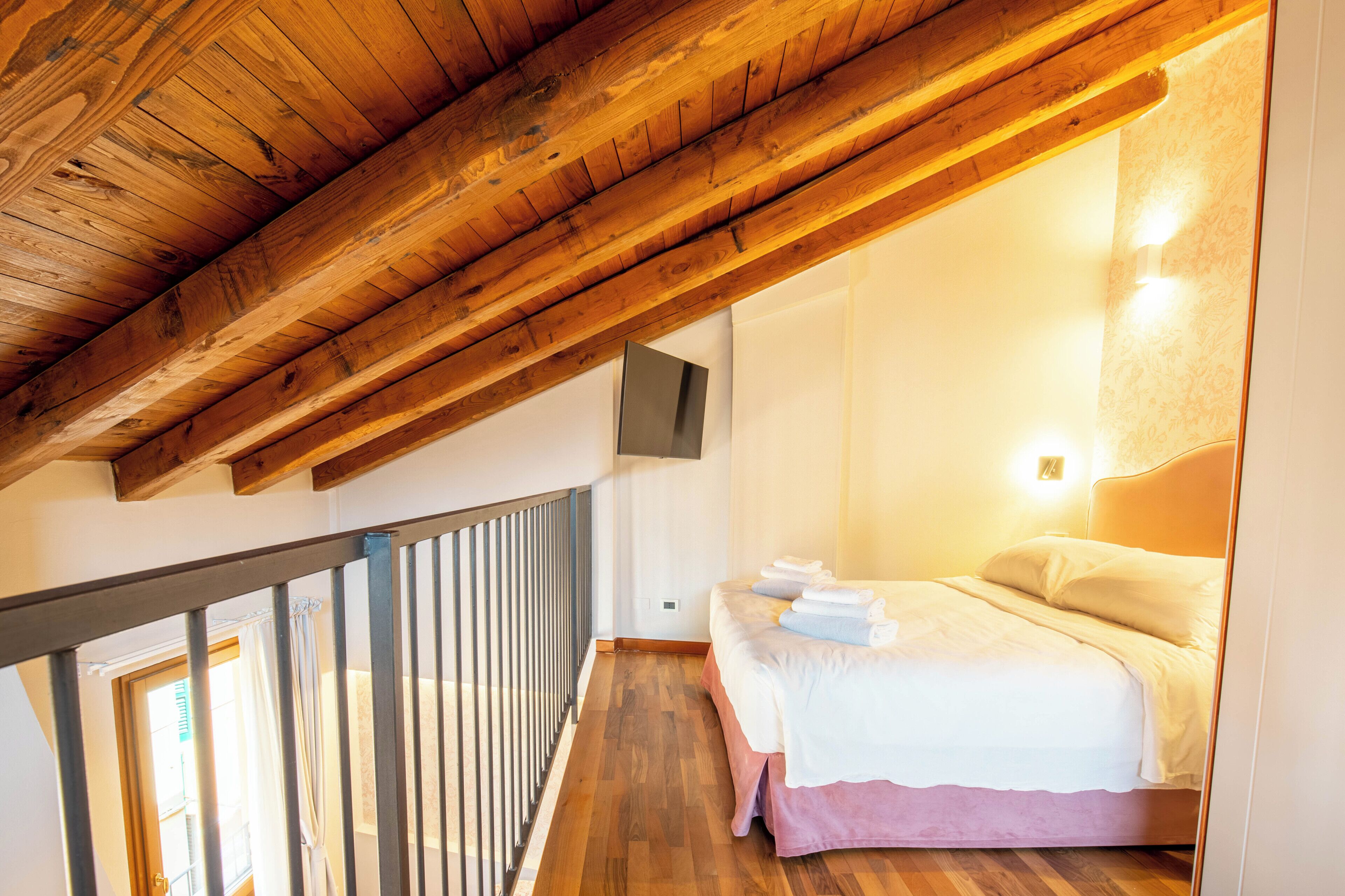 Une chambre avec un plafond en bois, un lit recouvert de draps blancs, un téléviseur fixé au mur et une fenêtre avec des rideaux.
