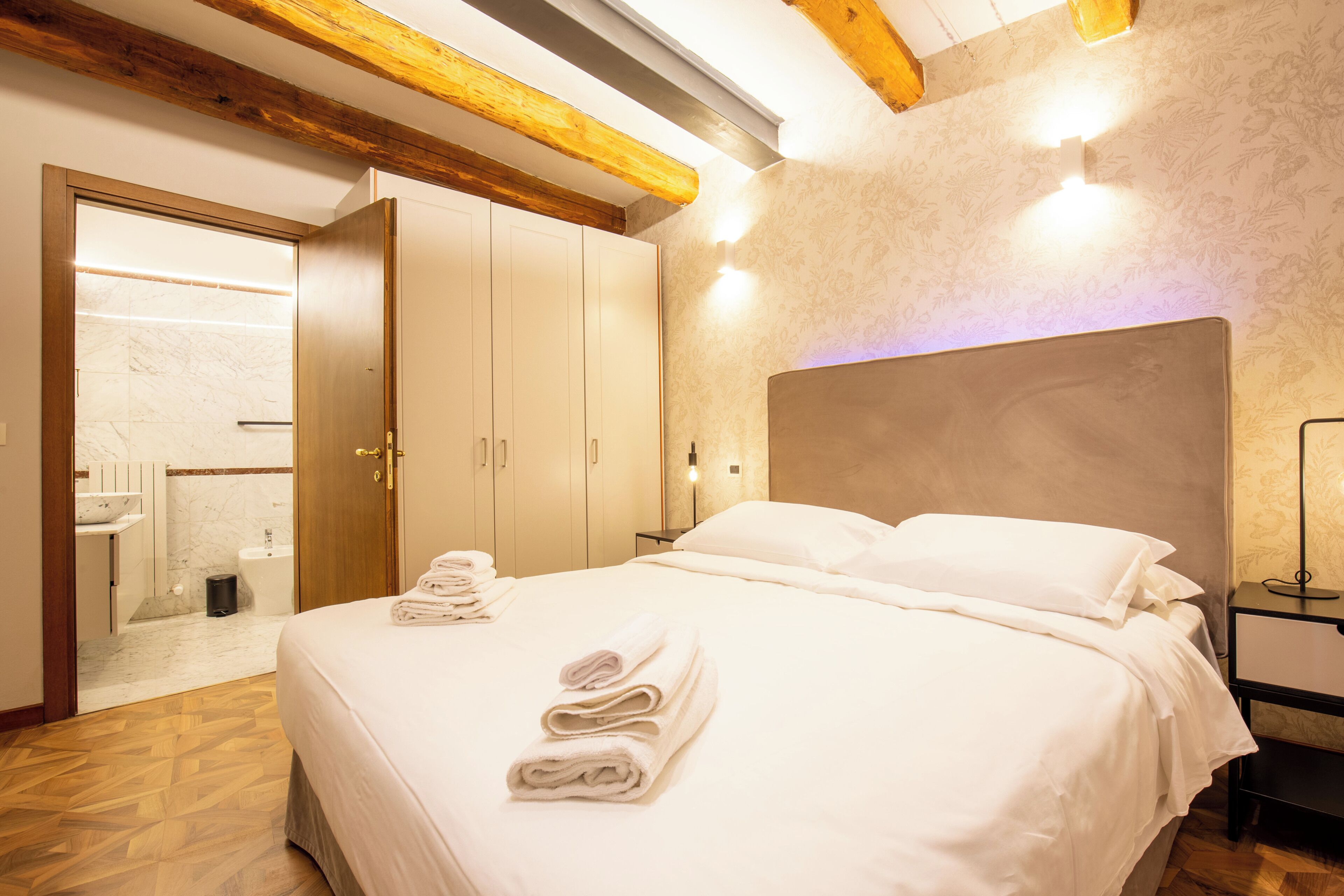A hotel room with a large bed, a wooden ceiling, a bathroom with a marble sink, and a nightstand with a lamp.