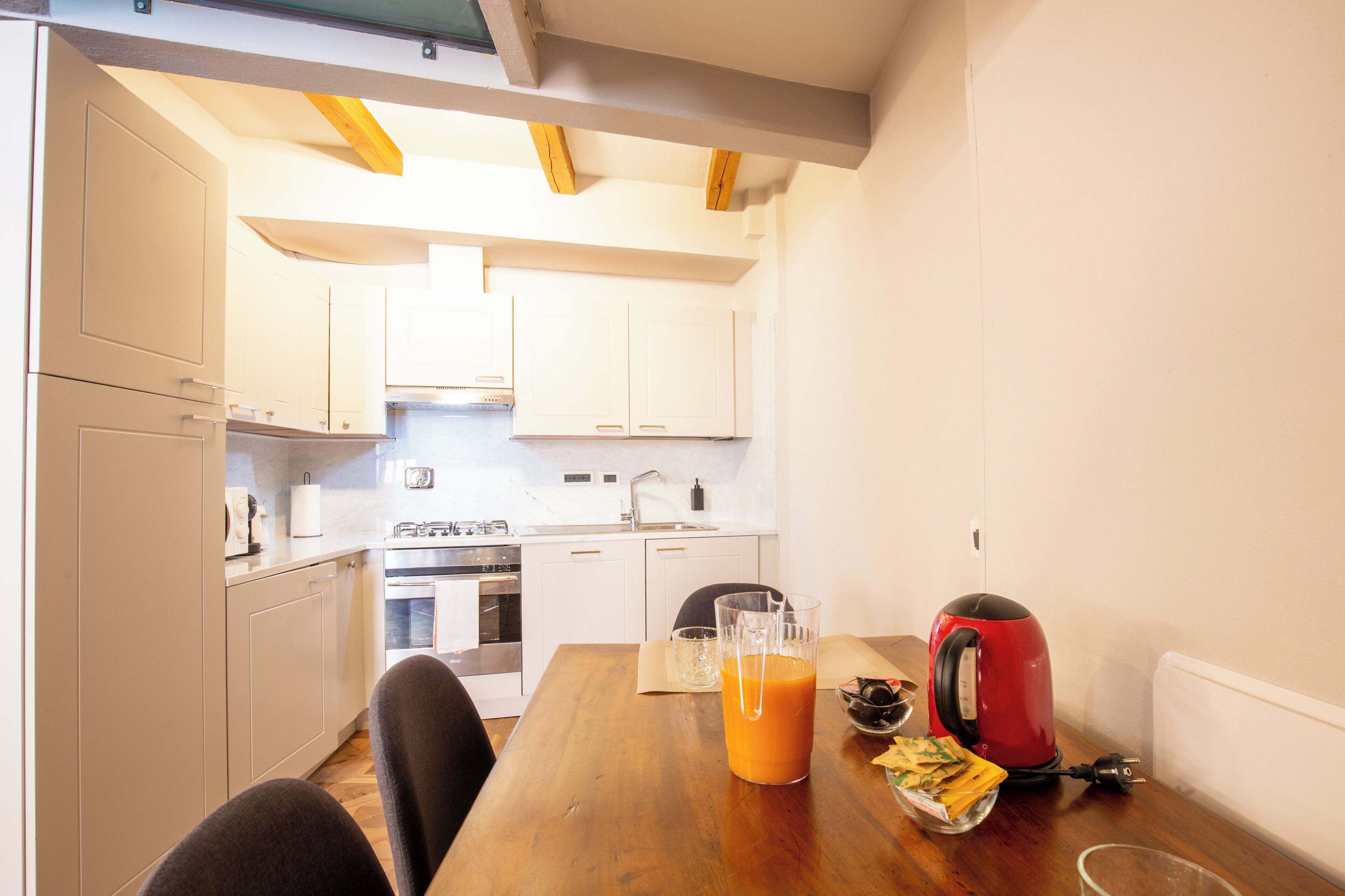 Una cocina con armarios blancos, una mesa de madera, un hervidor rojo y un vaso de jugo de naranja.