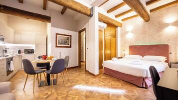 A hotel room with a wooden ceiling, a bed with white linens, a dining table with chairs, and a kitchenette.