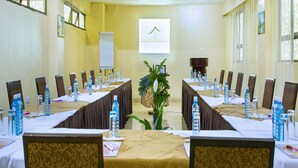Un salón de conferencias con largas mesas, sillas, botellas de agua y una pantalla de proyector.