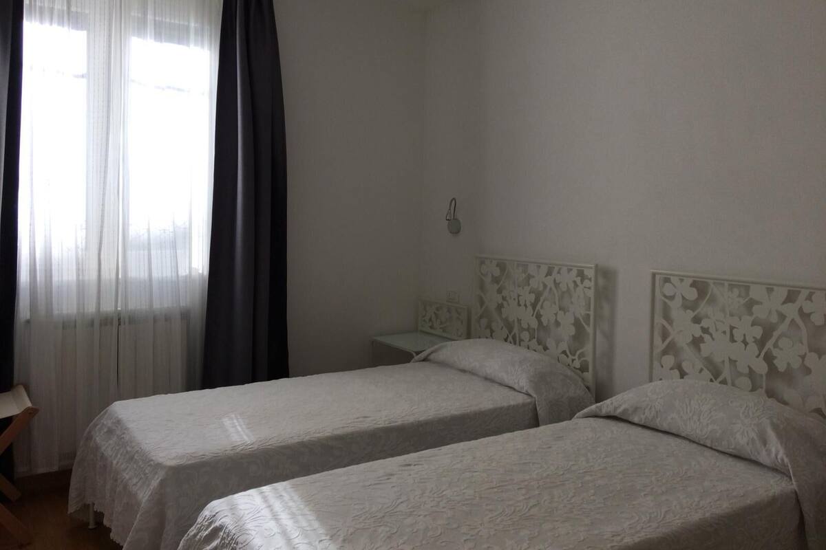 A room with two single beds, a small bedside table, a wall-mounted light, and a window with sheer curtains and a dark drape.