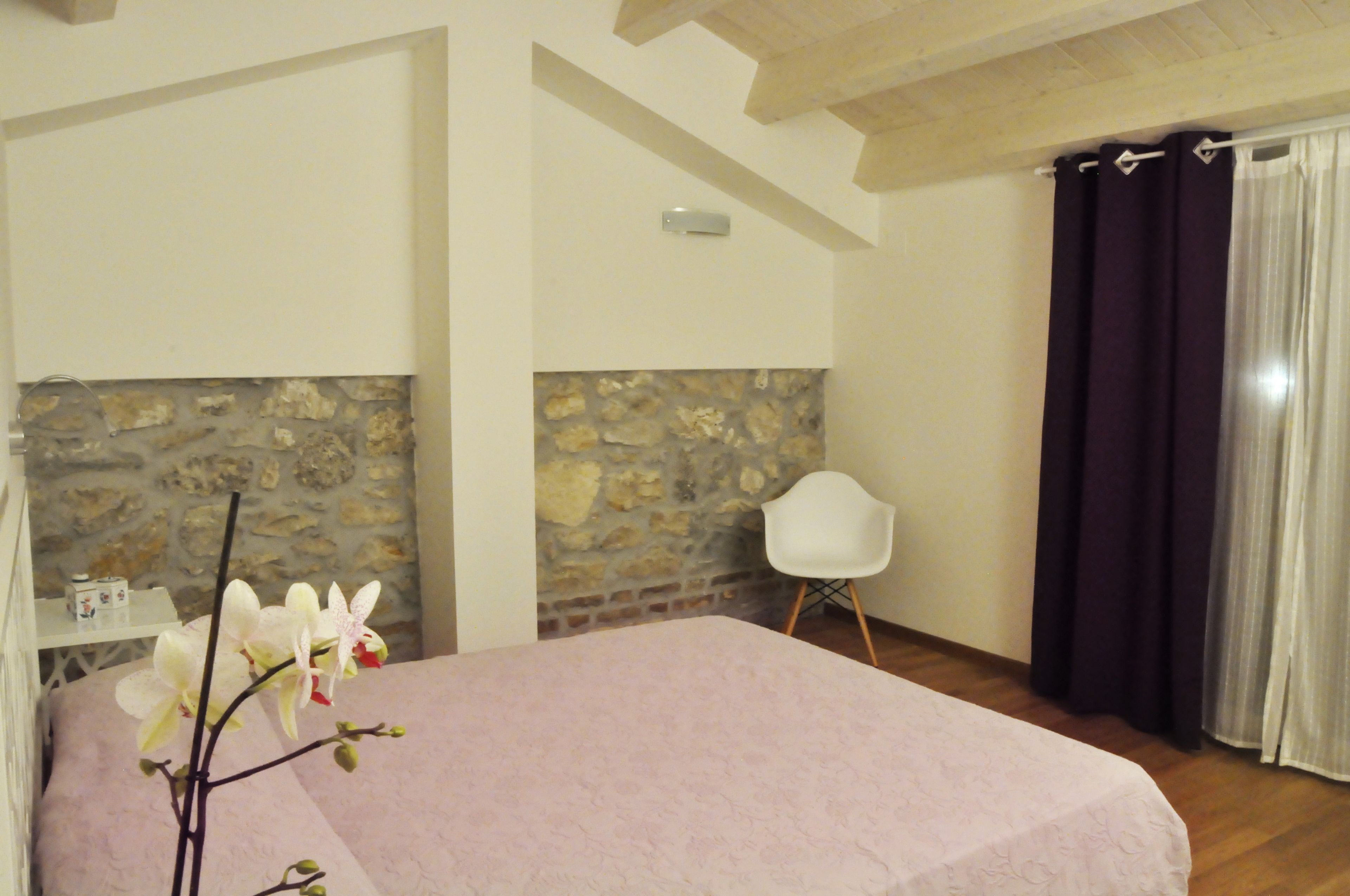 Een slaapkamer met een stenen muur, een bed met een roze dekbedovertrek, een witte stoel en een raam met donkere gordijnen.