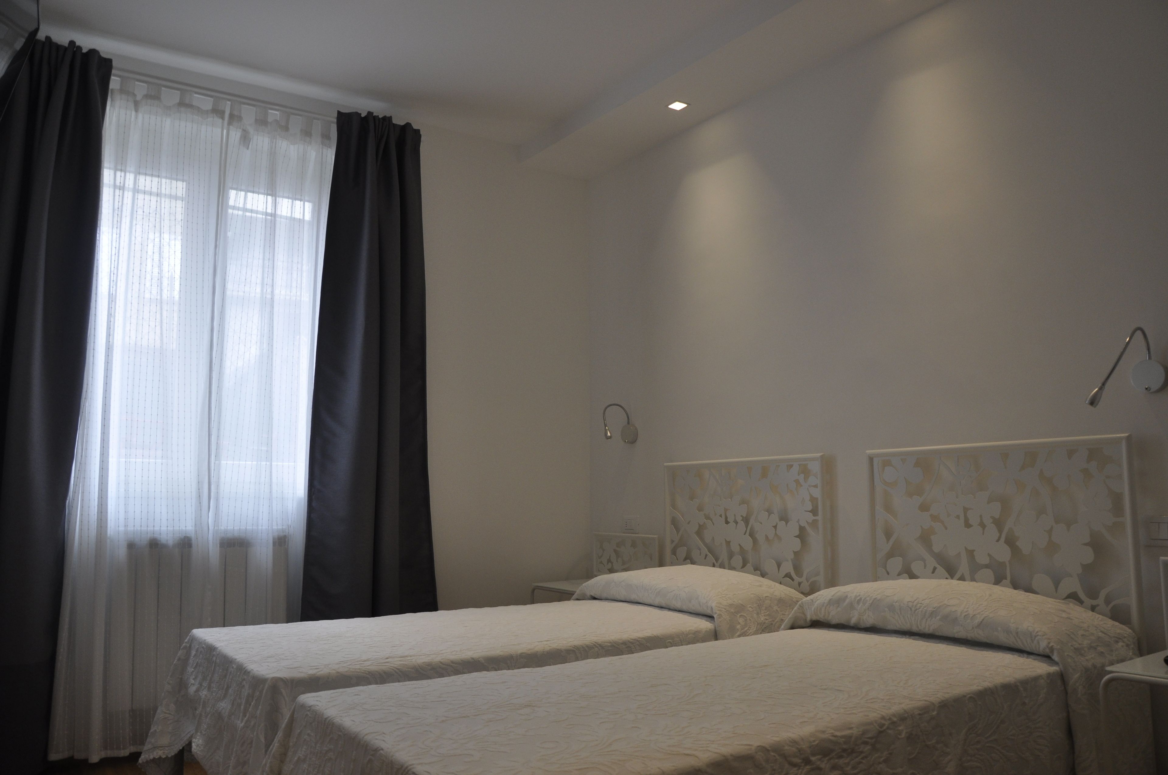 A bedroom with two beds, a window with sheer curtains, and a wall-mounted light fixture.