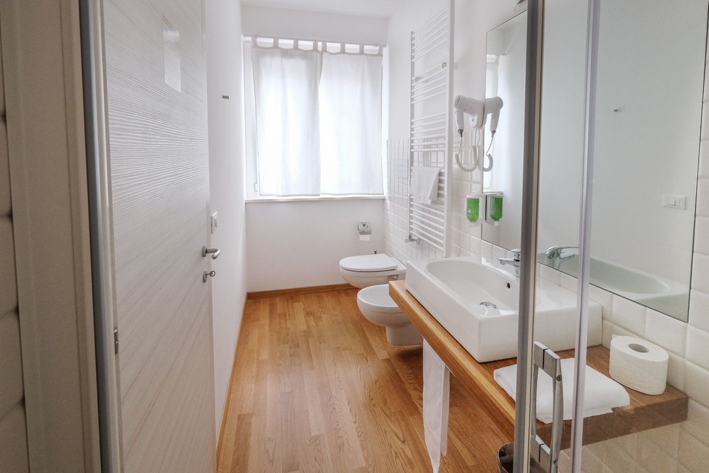 Un baño moderno con suelo de madera, sanitarios blancos, cabina de ducha de cristal y un espejo grande.